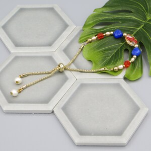 May include: A gold chain bracelet with red, blue and white beads. The bracelet has a sliding clasp and is displayed on a grey hexagonal tile.