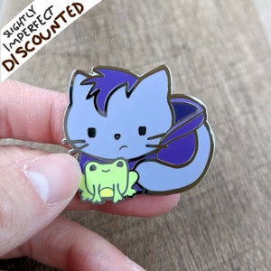 May include: Purple and silver enamel pin featuring a cartoon cat holding a green frog. The cat has a sad expression and is wearing a purple cape.