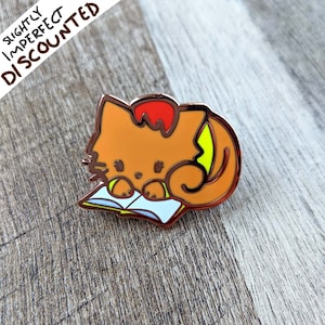 May include: Orange enamel pin of a cat reading a book. The cat has a red head and a yellow tail. The book is white with a blue cover.