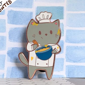 Può includere: Un gatto grigio con un cappello da cuoco bianco e un grembiule sta mescolando l'impasto in una ciotola blu. Il gatto ha schizzi di vernice gialla sul pelo.