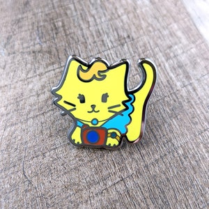 May include: A yellow enamel pin of a cat wearing a blue shirt and holding a camera. The cat has a yellow and orange swirl on its head.