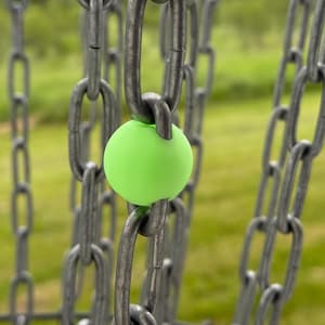 May include: A close-up of a disc golf basket chain with a bright green sphere. The chains are made of metal and are gray. The sphere is a vibrant green color and is positioned in the center of the chains. The background is blurred green grass.