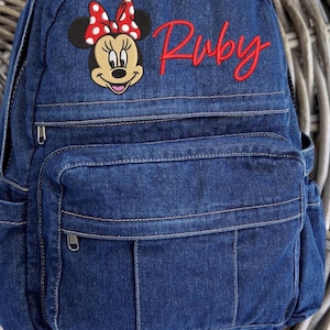 Pode incluir: Uma mochila de ganga azul-escura com um emblema bordado da Minnie Mouse com um la&ccedil;o vermelho de bolinhas brancas. O nome Ruby est&aacute; bordado em letra cursiva vermelha no lado superior direito. A mochila tem uma al&ccedil;a superior e um bolso frontal.