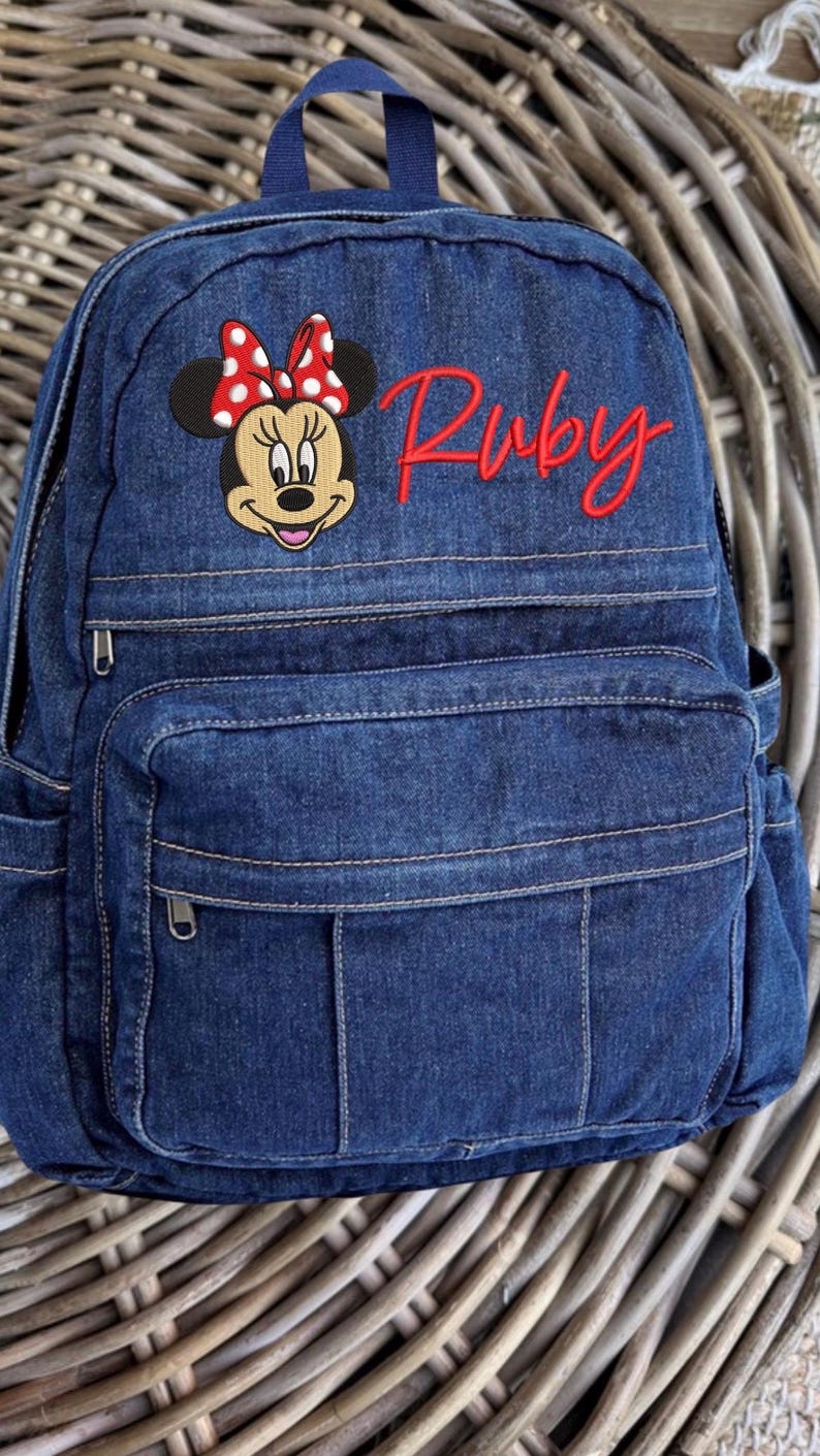 Pode incluir: Uma mochila de ganga azul-escura com um emblema bordado da Minnie Mouse com um la&ccedil;o vermelho de bolinhas brancas. O nome Ruby est&aacute; bordado em letra cursiva vermelha no lado superior direito. A mochila tem uma al&ccedil;a superior e um bolso frontal.