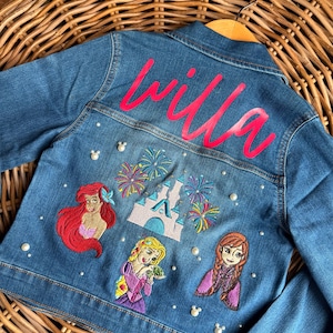 May include: A blue denim jacket with embroidered Disney princesses, a castle, and fireworks. The jacket has pearl-like embellishments and the name "Willa" is embroidered on the back in pink.