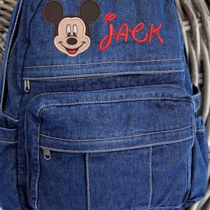 May include: A dark blue denim backpack featuring an embroidered Mickey Mouse head and the name Jack stitched in red thread. The bag includes a top handle, a main zippered compartment, and a smaller front zippered pocket with vertical stitching details.