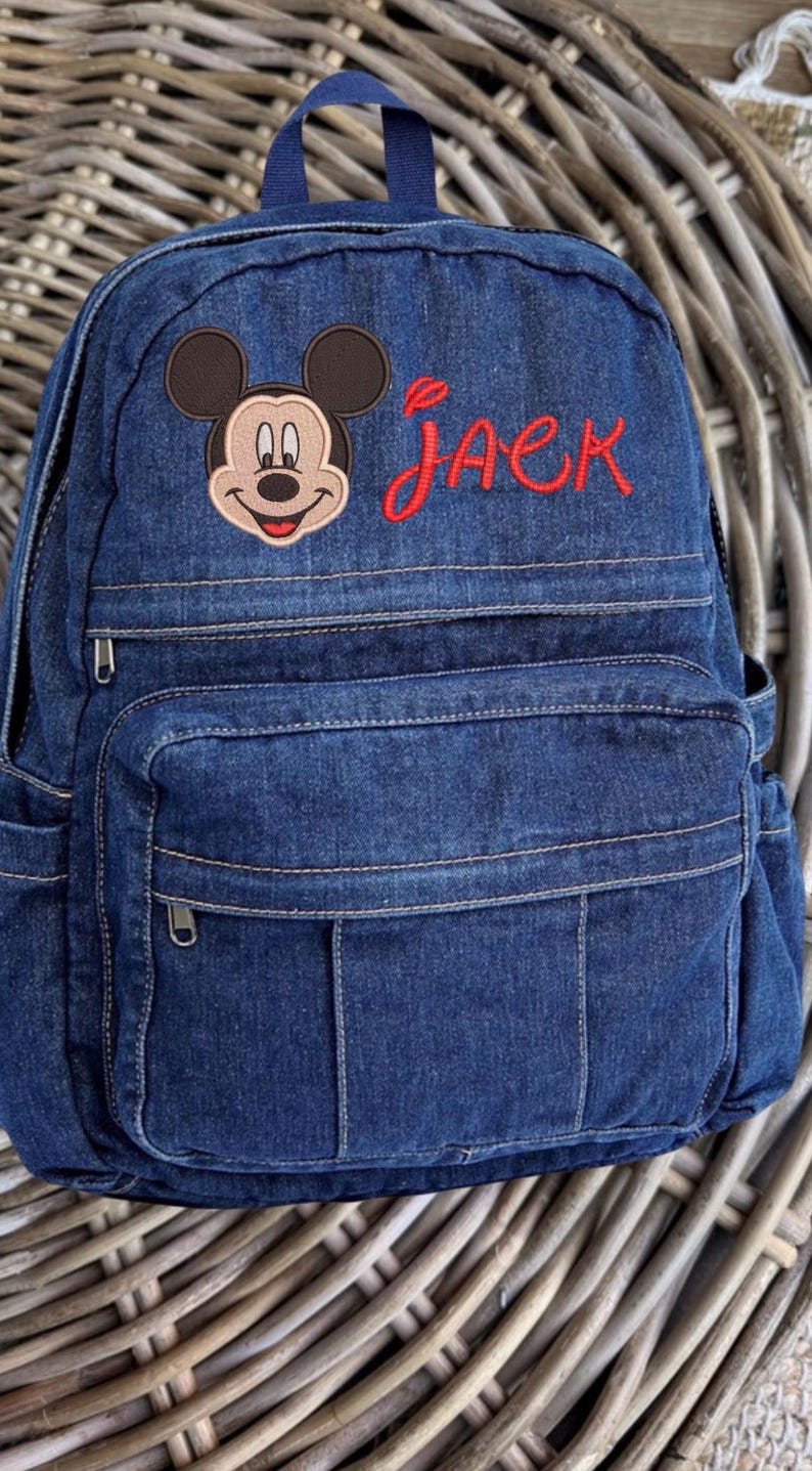 May include: A dark blue denim backpack featuring an embroidered Mickey Mouse head and the name Jack stitched in red thread. The bag includes a top handle, a main zippered compartment, and a smaller front zippered pocket with vertical stitching details.