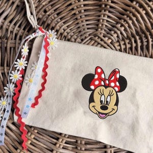 May include: A beige canvas pouch with an embroidered Minnie Mouse face and a red polka dot bow. The pouch has a cream strap and is decorated with daisy and star ribbons. The background is a woven wicker basket.