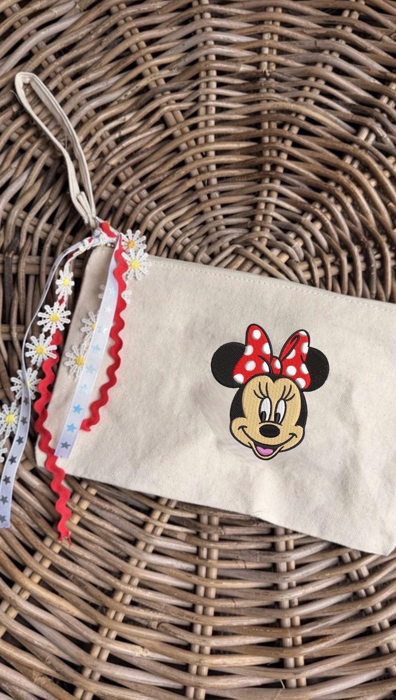 May include: A beige canvas pouch with an embroidered Minnie Mouse face and a red polka dot bow. The pouch has a cream strap and is decorated with daisy and star ribbons. The background is a woven wicker basket.