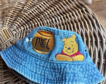 Könnte beinhalten: Ein blauer Jeans-Bucket-Hut mit gesticktem Winnie Puuh und einem Honigtopf mit dem Wort "MEL" darauf.