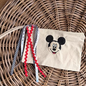 Pode incluir: Bolsa de lona bege com um rosto de Mickey Mouse bordado. A bolsa tem uma al&ccedil;a longa e &eacute; decorada com fitas vermelhas, pretas e brancas. A bolsa est&aacute; sobre uma superf&iacute;cie de vime tecido.