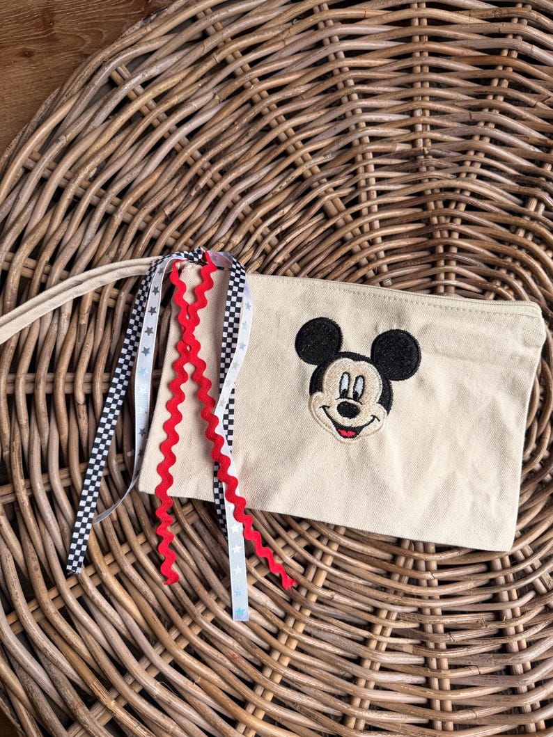 Pode incluir: Bolsa de lona bege com um rosto de Mickey Mouse bordado. A bolsa tem uma al&ccedil;a longa e &eacute; decorada com fitas vermelhas, pretas e brancas. A bolsa est&aacute; sobre uma superf&iacute;cie de vime tecido.
