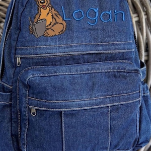May include: A dark blue denim backpack featuring a top handle, a front zippered pocket, and side pockets. The front is embroidered with a brown bear holding a grey book and the name Logan in blue thread, showcasing a classic fabric texture.