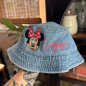 May include: A light blue denim bucket hat featuring an embroidered patch of Minnie Mouse with a pink polka dot bow. The name Logan is embroidered in pink cursive script on the side of the hat. The hat has a wide, downward-sloping brim.