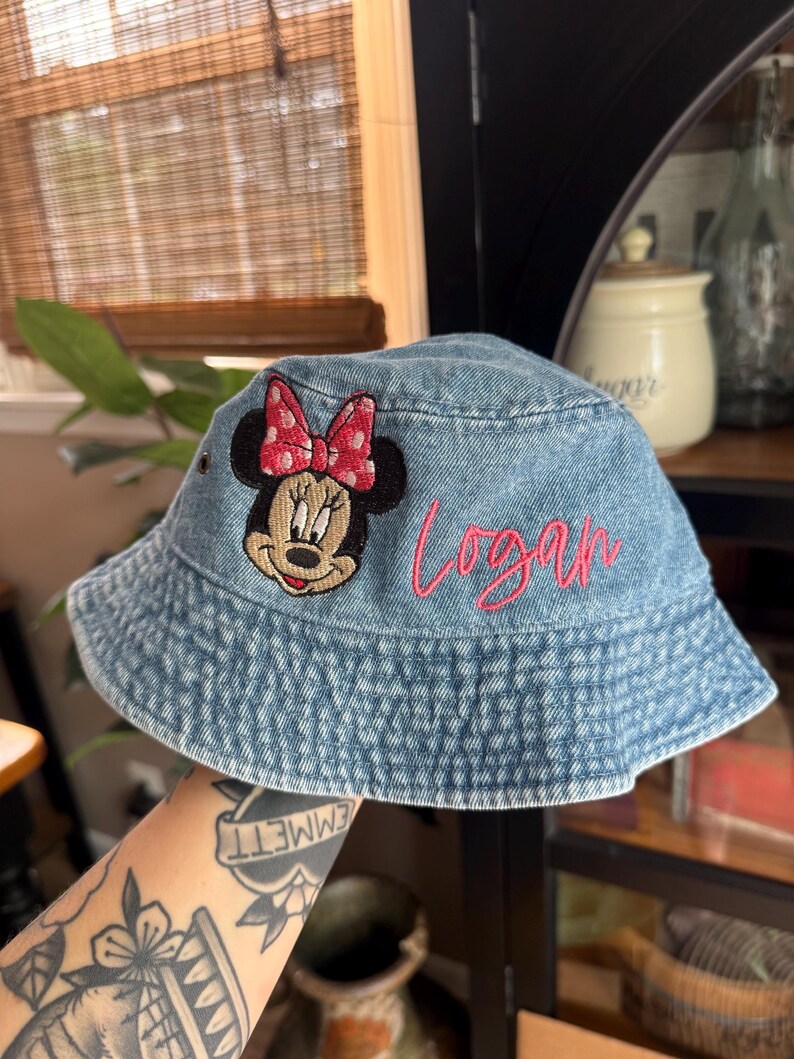 May include: A light blue denim bucket hat featuring an embroidered patch of Minnie Mouse with a pink polka dot bow. The name Logan is embroidered in pink cursive script on the side of the hat. The hat has a wide, downward-sloping brim.