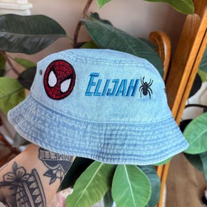 May include: Light blue denim bucket hat featuring a Spider-Man emblem and the name "Elijah" embroidered in blue. A black spider graphic is embroidered next to the name. The hat is a casual accessory.