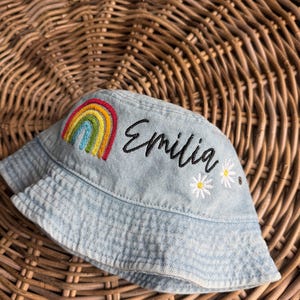 May include: A light blue denim bucket hat with a rainbow and the name "Emilia" embroidered on the front. The hat has white daisy accents.
