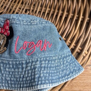 May include: A light blue denim bucket hat featuring an embroidered pink script name Logan and a partial embroidered character patch with a red polka dot bow. The hat includes a small metal grommet on the side for ventilation and stitching.