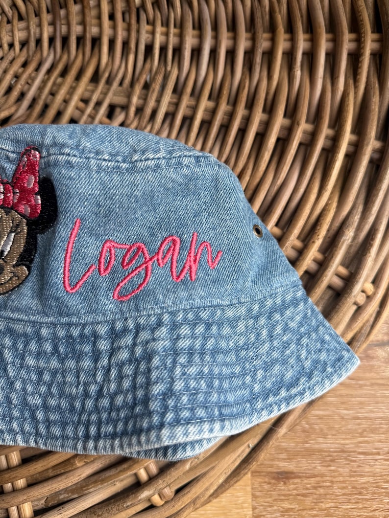 May include: A light blue denim bucket hat featuring an embroidered pink script name Logan and a partial embroidered character patch with a red polka dot bow. The hat includes a small metal grommet on the side for ventilation and stitching.