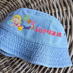 May include: A light blue denim bucket hat featuring an embroidered cartoon character with blonde hair lying down, surrounded by colorful flower icons in blue, green, pink, and yellow. The name Jordan is embroidered in red cursive script.