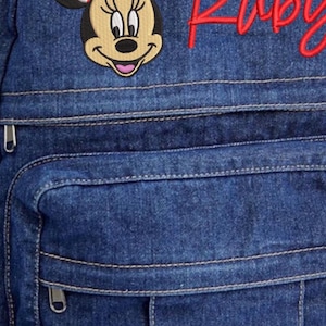 Pode incluir: Uma mochila de ganga azul com um emblema bordado da Minnie Mouse com um la&ccedil;o vermelho de bolinhas brancas. O nome Ruby est&aacute; bordado em letra cursiva vermelha no lado superior direito. A mala inclui um bolso frontal com fecho e al&ccedil;a.