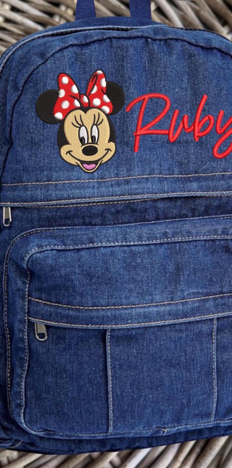 Pode incluir: Uma mochila de ganga azul com um emblema bordado da Minnie Mouse com um la&ccedil;o vermelho de bolinhas brancas. O nome Ruby est&aacute; bordado em letra cursiva vermelha no lado superior direito. A mala inclui um bolso frontal com fecho e al&ccedil;a.