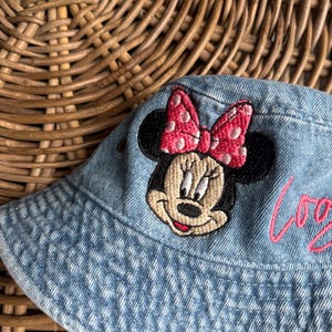May include: A light blue denim bucket hat featuring an embroidered patch of Minnie Mouse with a pink polka dot bow. The hat includes a small metal eyelet on the side and pink cursive text partially visible on the right side of the fabric.