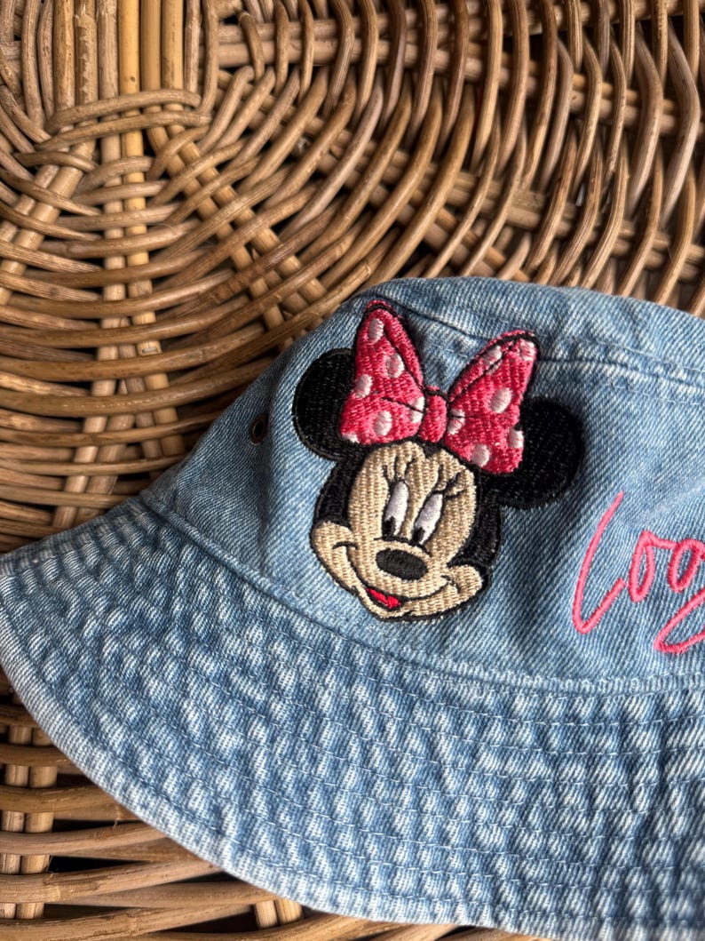 May include: A light blue denim bucket hat featuring an embroidered patch of Minnie Mouse with a pink polka dot bow. The hat includes a small metal eyelet on the side and pink cursive text partially visible on the right side of the fabric.
