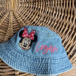 May include: A light blue denim bucket hat featuring an embroidered patch of Minnie Mouse with a pink polka dot bow and the name Logan stitched in pink cursive thread on the front of the crown. The hat has a downward sloping brim.