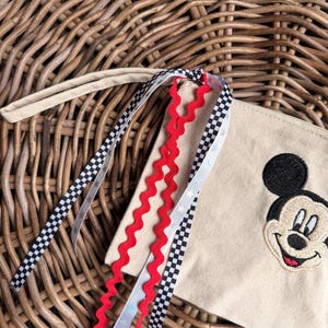 Pode incluir: Uma bolsa de lona bege com um rosto de Mickey Mouse bordado em preto. A bolsa tem uma al&ccedil;a de pulso e &eacute; decorada com fitas vermelhas, pretas e brancas. As fitas t&ecirc;m um padr&atilde;o xadrez e detalhes de estrelas.