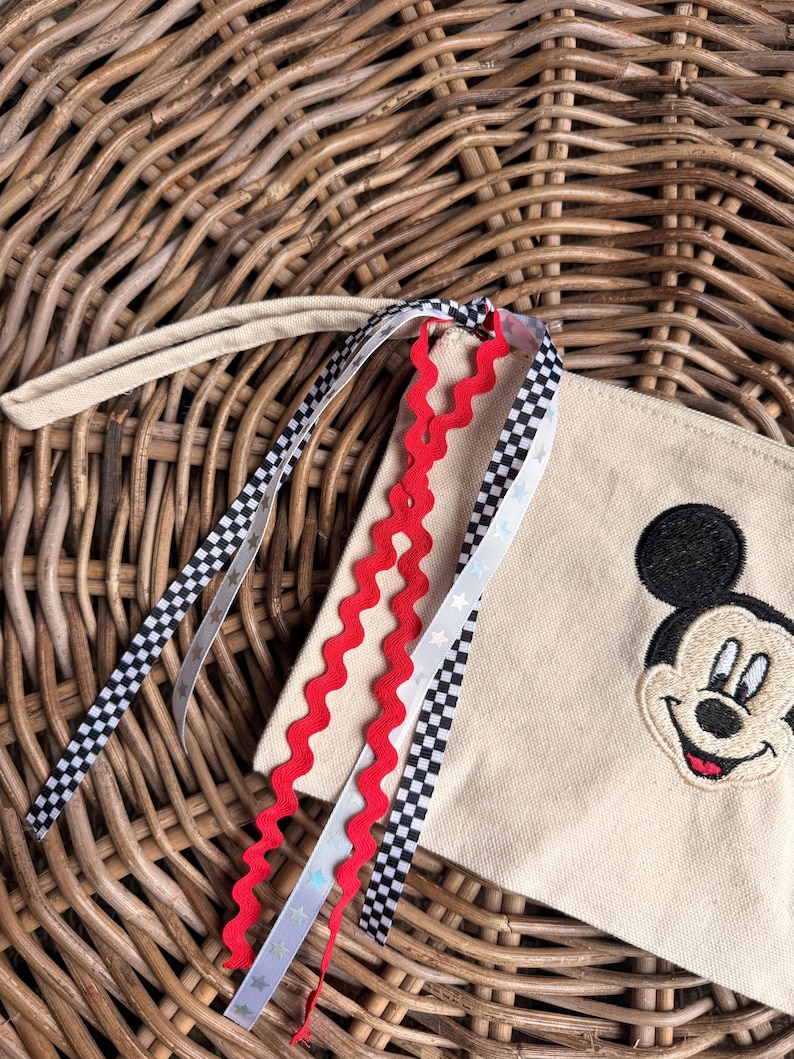 Pode incluir: Uma bolsa de lona bege com um rosto de Mickey Mouse bordado em preto. A bolsa tem uma al&ccedil;a de pulso e &eacute; decorada com fitas vermelhas, pretas e brancas. As fitas t&ecirc;m um padr&atilde;o xadrez e detalhes de estrelas.