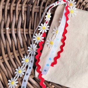 May include: Close-up of a beige canvas bag with decorative ribbons. The ribbons include white daisy chains, red zigzag trim, and a white ribbon with blue stars. The background is a woven wicker basket.