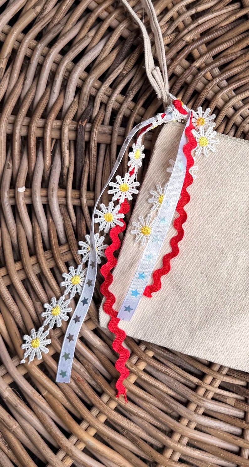May include: Close-up of a beige canvas bag with decorative ribbons. The ribbons include white daisy chains, red zigzag trim, and a white ribbon with blue stars. The background is a woven wicker basket.