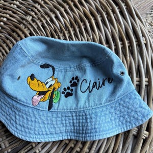May include: Light blue denim bucket hat featuring an embroidered Pluto cartoon character with a yellow head, black ears, and a pink tongue. The hat also has black paw prints and the name "Claire" in cursive.