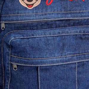 May include: A dark blue denim backpack featuring an embroidered Mickey Mouse head and the name JACK stitched in red thread. The backpack includes a front zippered pocket with visible stitching details and silver-toned metal zipper pulls.