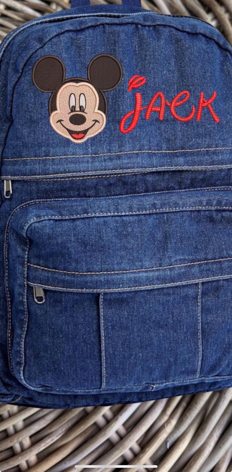 May include: A dark blue denim backpack featuring an embroidered Mickey Mouse head and the name JACK stitched in red thread. The backpack includes a front zippered pocket with visible stitching details and silver-toned metal zipper pulls.