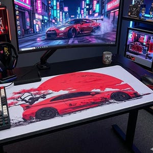 May include: A rectangular desk mat featuring a large red sun graphic and a red sports car illustration. The mat is positioned on a black desk surface next to a mechanical keyboard, a computer mouse, a gaming headset, and a monitor displaying a similar car.
