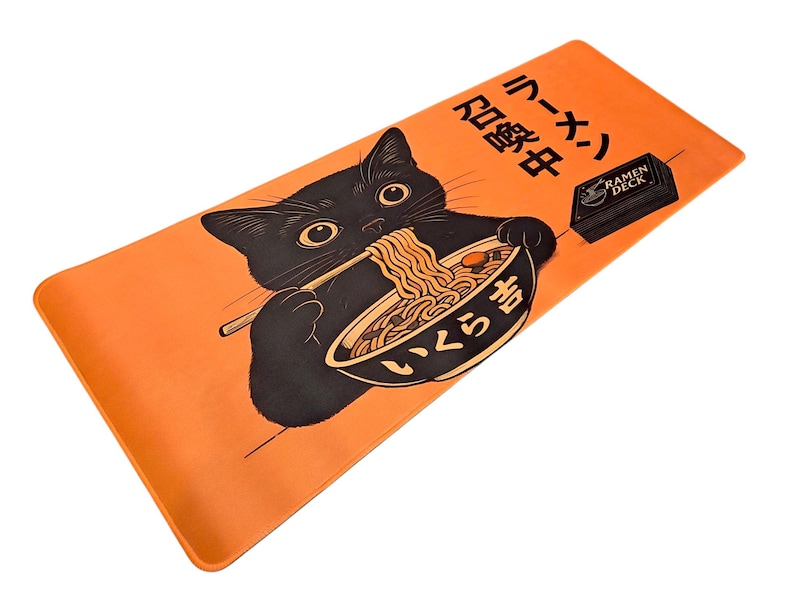 May include: A rectangular orange desk mat featuring a black cat illustration eating ramen with chopsticks from a bowl. The text includes Japanese characters and the words RAMEN DECK printed in black. The design is centered on the vibrant orange surface.
