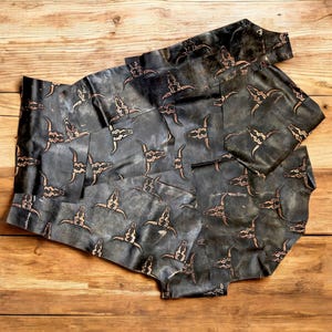 May include: Dark grey leather pieces featuring a repeating pattern of tan bull skulls. The leather has a distressed, textured appearance, and the skulls are evenly spaced. The pieces are arranged on a wooden surface.