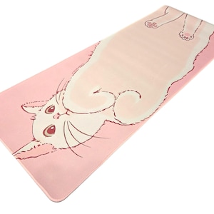 May include: A rectangular yoga mat featuring a light pink background with a large, cream-colored cat illustration stretched across the surface. The cat has dark pink outlines, large eyes, and visible paw pads, extending from the bottom left to the top right.