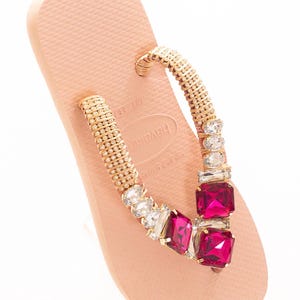 May include: A light pink Havaianas flip-flop with a gold beaded strap. The strap is decorated with clear and fuchsia-coloured square and rectangular gemstones. The Havaianas logo is visible on the sole.