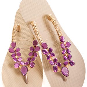 以下が含まれることがあります： ベージュ色の Havaianas ビーチサンダル。ストラップは、金色のビーズと紫色の宝石のような花のクラスターで飾られています。ビーチサンダルは白い背景に置かれています。
