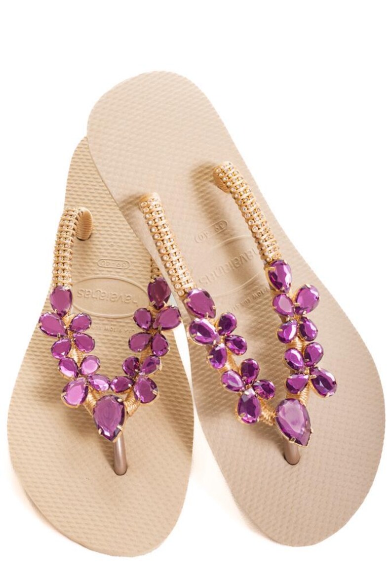 以下が含まれることがあります： ベージュ色の Havaianas ビーチサンダル。ストラップは、金色のビーズと紫色の宝石のような花のクラスターで飾られています。ビーチサンダルは白い背景に置かれています。