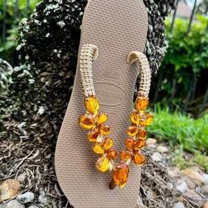 Pode incluir: Um par de chinelos marrons com uma tira de contas douradas adornadas com contas de cor &acirc;mbar. A tira &eacute; decorada com pequenos strass.