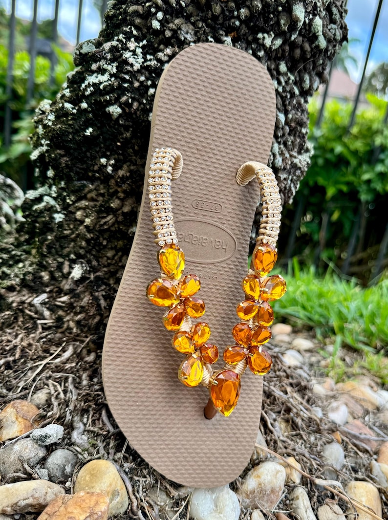 Pode incluir: Um par de chinelos marrons com uma tira de contas douradas adornadas com contas de cor &acirc;mbar. A tira &eacute; decorada com pequenos strass.