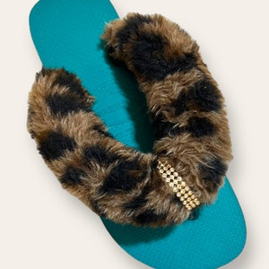 May include: Teal flip flops with a brown and black leopard print fur strap and a gold rhinestone embellishment.