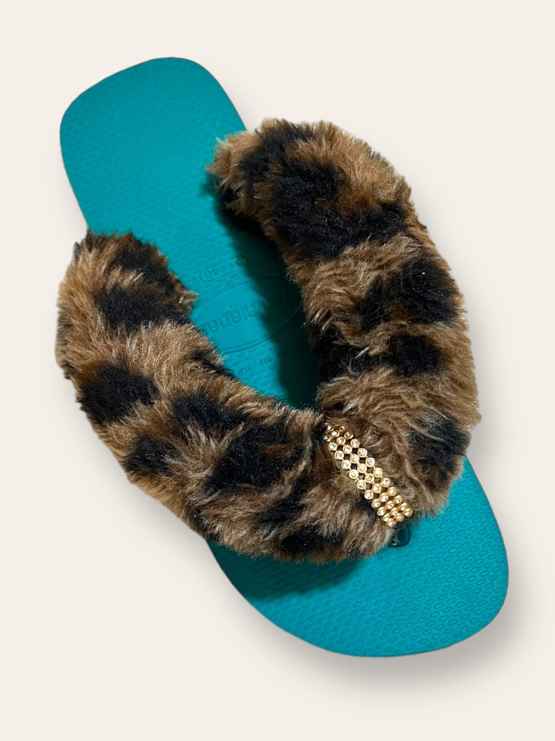 May include: Teal flip flops with a brown and black leopard print fur strap and a gold rhinestone embellishment.