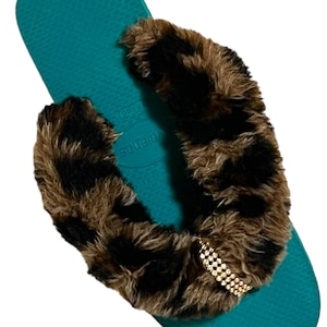 May include: Teal flip flops with a brown and black faux fur strap and a gold rhinestone embellishment. The strap says "Havaianas" in white.