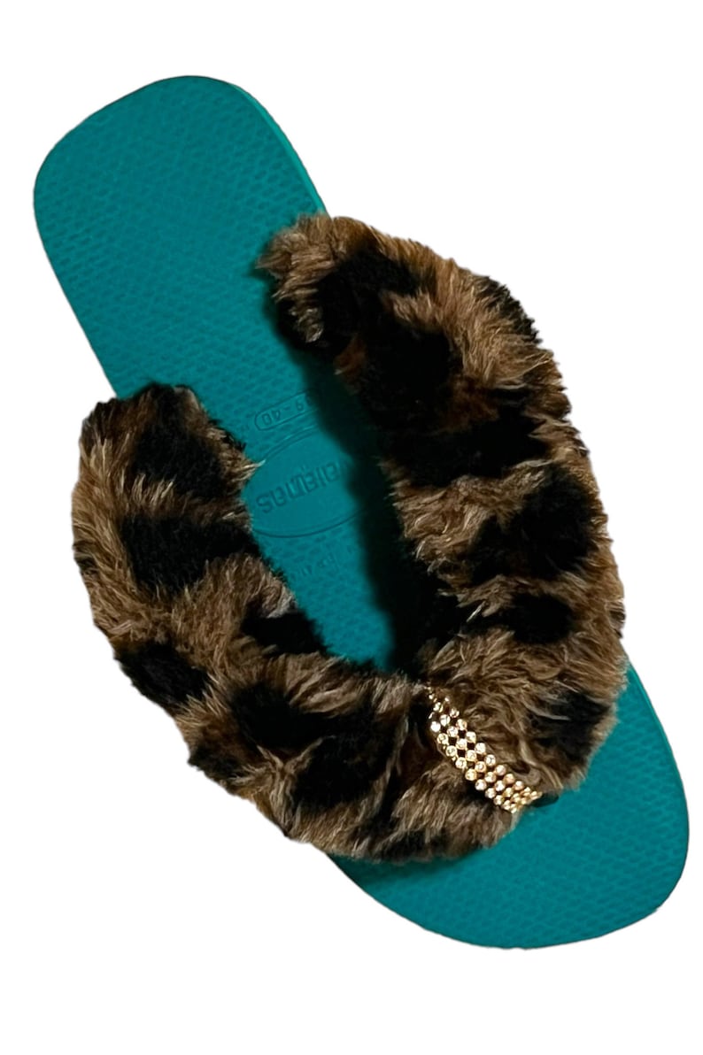 May include: Teal flip flops with a brown and black faux fur strap and a gold rhinestone embellishment. The strap says "Havaianas" in white.