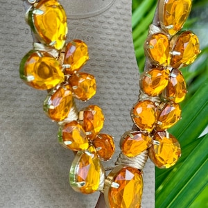 Pode incluir: Um par de chinelos marrons com uma tira dourada adornada com cristais laranja. A tira apresenta um design floral com m&uacute;ltiplos cristais.